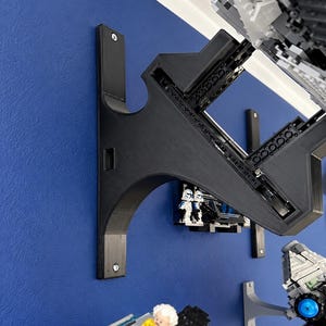 K&ouml;nnte beinhalten: Eine schwarze, 3D-gedruckte Wandhalterung mit einem geschwungenen St&uuml;tzarm und einer rechteckigen Grundplatte, die mit zwei Schrauben befestigt ist. Die Halterung dient zur Aufnahme eines gro&szlig;en grauen Kunststoff-Modellbausatzes mit sichtbarem Rahmen.