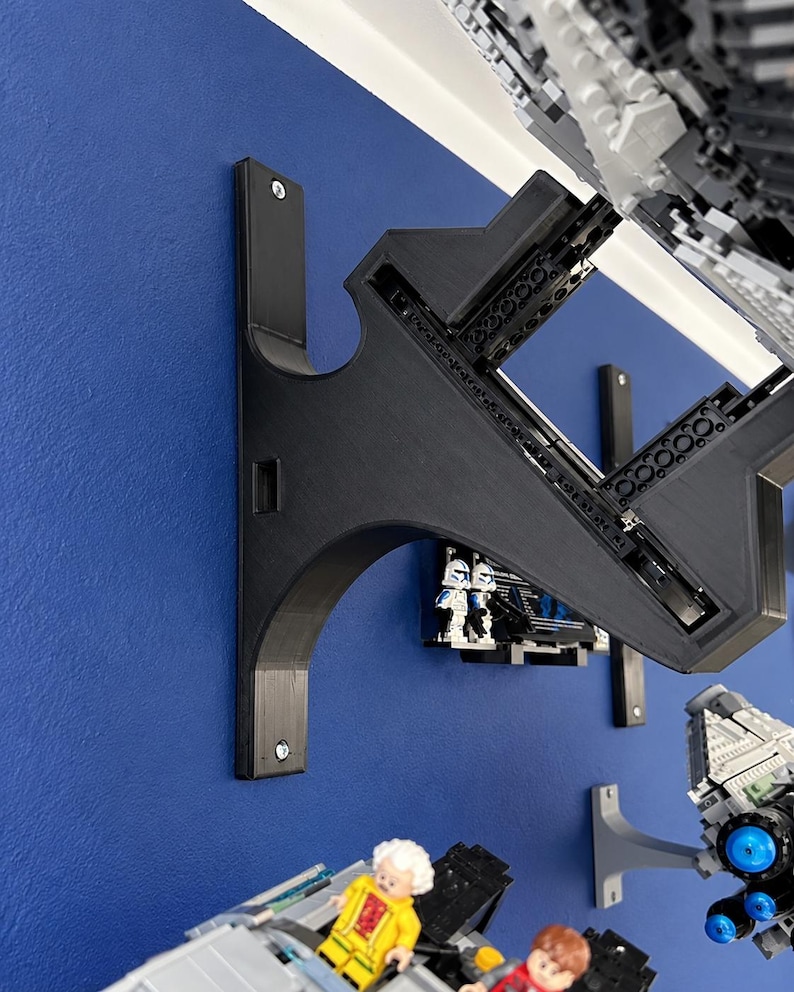 K&ouml;nnte beinhalten: Eine schwarze, 3D-gedruckte Wandhalterung mit einem geschwungenen St&uuml;tzarm und einer rechteckigen Grundplatte, die mit zwei Schrauben befestigt ist. Die Halterung dient zur Aufnahme eines gro&szlig;en grauen Kunststoff-Modellbausatzes mit sichtbarem Rahmen.