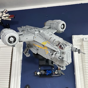 May include: A grey and yellow Lego Star Wars ship, the "Ghost", is mounted on a black stand against a blue wall. The ship has two engines and a cockpit.