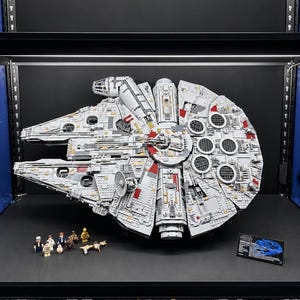 Pu&ograve; includere: Un grande e dettagliato modello LEGO Star Wars Millennium Falcon in grigio chiaro con accenti rossi, caratterizzato da uno scafo circolare, cabina di pilotaggio e parabola radar. Include diverse piccole minifigure e una targa informativa nera.