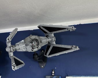 May include: A gray TIE Interceptor Lego model mounted on a black wall mount. The model is on a blue wall with a white trim. The model is in a room with a white ceiling.