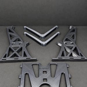 Pu&ograve; includere: Un set di componenti in plastica grigio scuro stampati in 3D che include due supporti a traliccio triangolari alti, due rinforzi angolari a forma di V e una piastra di base rettangolare con ritagli. La piastra di base presenta il testo "brimmingBricks."