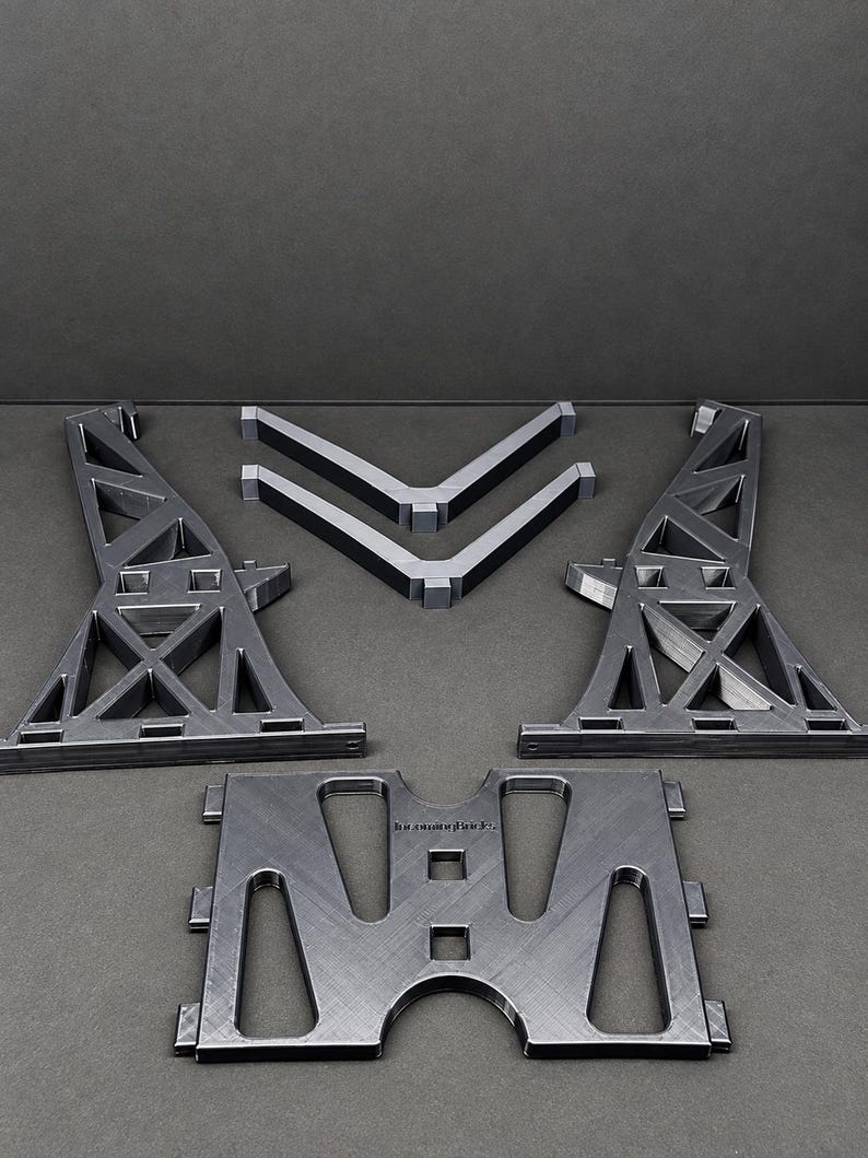 Pu&ograve; includere: Un set di componenti in plastica grigio scuro stampati in 3D che include due supporti a traliccio triangolari alti, due rinforzi angolari a forma di V e una piastra di base rettangolare con ritagli. La piastra di base presenta il testo "brimmingBricks."