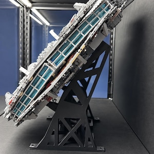 Pu&ograve; includere: Un grande e dettagliato modello di astronave grigia costruito con mattoncini di plastica a incastro, montato ad angolo su un supporto espositivo nero. La nave presenta un pannello laterale con file di elementi luminosi blu traslucidi.