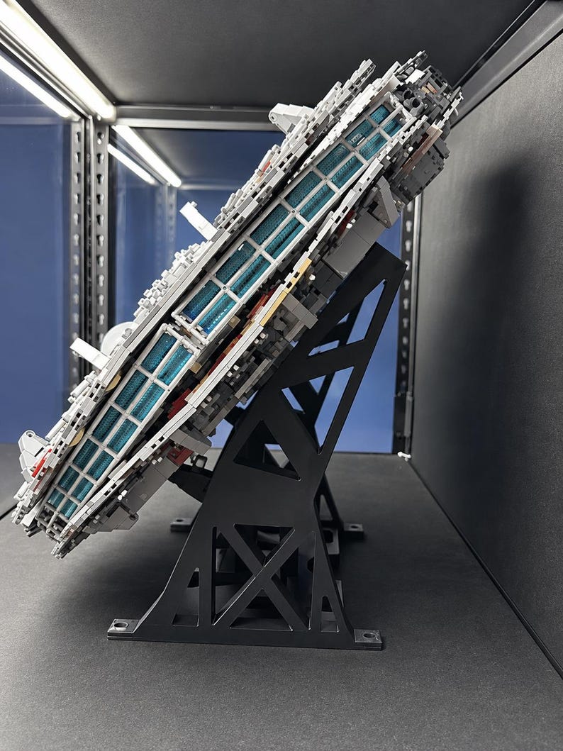 Pu&ograve; includere: Un grande e dettagliato modello di astronave grigia costruito con mattoncini di plastica a incastro, montato ad angolo su un supporto espositivo nero. La nave presenta un pannello laterale con file di elementi luminosi blu traslucidi.