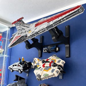 K&ouml;nnte beinhalten: Ein gro&szlig;es LEGO Star Wars Venator-class Republic Attack Cruiser Modell auf einer schwarzen Wandhalterung, positioniert &uuml;ber einem LEGO DeLorean, einem wei&szlig;-beigen Raumschiff, einem grauen Sternenj&auml;ger und einem orangefarbenen Raumschiff.