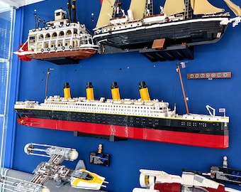 Könnte beinhalten: Eine Sammlung von aus Bausteinen gefertigten Modellen an einer blauen Wand, darunter ein weiß-schwarzer Raddampfer, ein großes schwarz-rotes Titanic-Schiff, ein Segelschiff mit beigen Segeln, ein grauer Y-Wing-Sternjäger und ein rot-weißes Raumschiff.