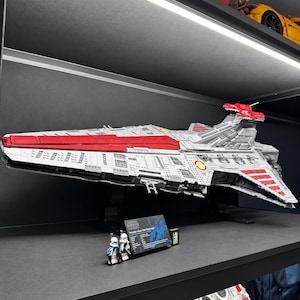 K&ouml;nnte beinhalten: Ein gro&szlig;es, detailliertes LEGO-Modell eines Venator-Klasse Sternenzerst&ouml;rers in Grau und Rot, montiert auf einem schwarzen St&auml;nder. Zwei kleine Klonkrieger-Minifiguren stehen neben einer schwarzen Infotafel mit wei&szlig;em Text zu den Schiffsdaten.
