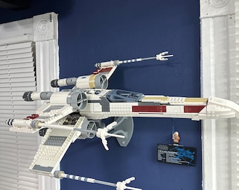 May include: A white, red, and gold LEGO Star Wars X-Wing Fighter model mounted on a wall. The model has two laser cannons and a pilot figure in the cockpit. The model is displayed with a small instruction booklet.