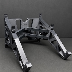 Pu&ograve; includere: Un supporto per laptop grigio scuro stampato in 3D, caratterizzato da un telaio geometrico a traliccio con aperture triangolari. La struttura include due bracci di supporto angolati con ganci frontali e un pannello posteriore con il testo IncommOptics.