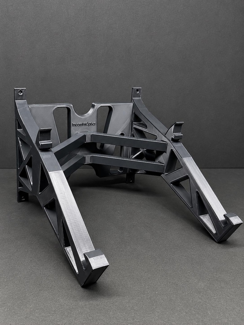 Pu&ograve; includere: Un supporto per laptop grigio scuro stampato in 3D, caratterizzato da un telaio geometrico a traliccio con aperture triangolari. La struttura include due bracci di supporto angolati con ganci frontali e un pannello posteriore con il testo IncommOptics.