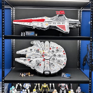 K&ouml;nnte beinhalten: Ein schwarzes Metallregal mit einem roten und einem gelben Sportwagen auf der obersten Ebene, einem gro&szlig;en grauen Raumschiffmodell mit zwei Minifiguren auf der zweiten, einem runden grauen Raumschiffmodell mit mehreren Minifiguren auf der dritten und verschiedenen Actionfiguren auf der untersten Ebene.