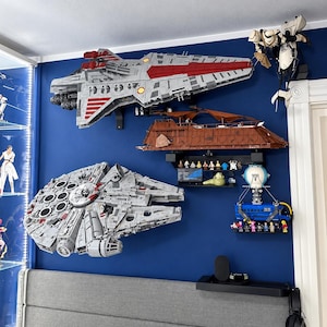 K&ouml;nnte beinhalten: Eine Sammlung von Star Wars-Modellen aus Bausteinen an einer blauen Wand, darunter ein gro&szlig;es graues Raumschiff, ein brauner Sandcrawler, ein kreisf&ouml;rmiger grauer Frachter, ein blaues Transportfahrzeug und verschiedene kleine Figuren.