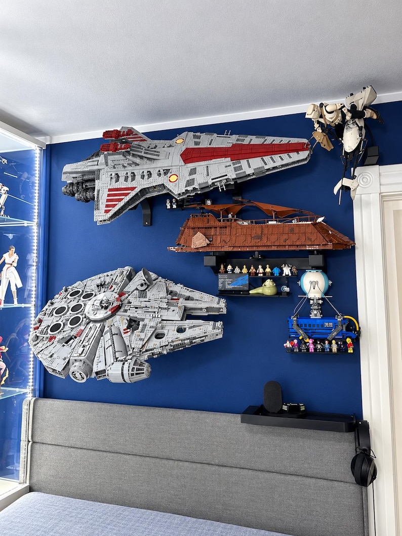 K&ouml;nnte beinhalten: Eine Sammlung von Star Wars-Modellen aus Bausteinen an einer blauen Wand, darunter ein gro&szlig;es graues Raumschiff, ein brauner Sandcrawler, ein kreisf&ouml;rmiger grauer Frachter, ein blaues Transportfahrzeug und verschiedene kleine Figuren.