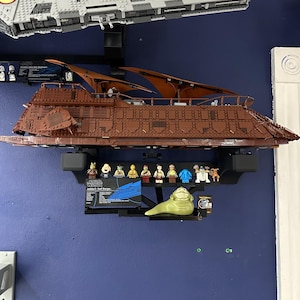 Könnte beinhalten: Ein braunes und beiges Lego Star Wars Jabba's Sail Barge mit einem roten und grauen Lego Star Wars Raumschiff im Hintergrund. Das Schiff ist auf einem Wandregal mit einer Vielzahl von Lego Star Wars Minifiguren und einer Jabba the Hutt Figur ausgestellt.