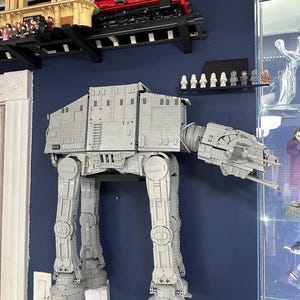 Könnte beinhalten: Ein großes, graues AT-AT-Modell aus Star Wars, konstruiert aus ineinandergreifenden Bausteinen, an einer dunkelblauen Wand befestigt. Darüber sind ein rot-schwarzes Zugmodell und ein Gebäudemodell ausgestellt. Mehrere kleine Figuren sind ebenfalls zu sehen.