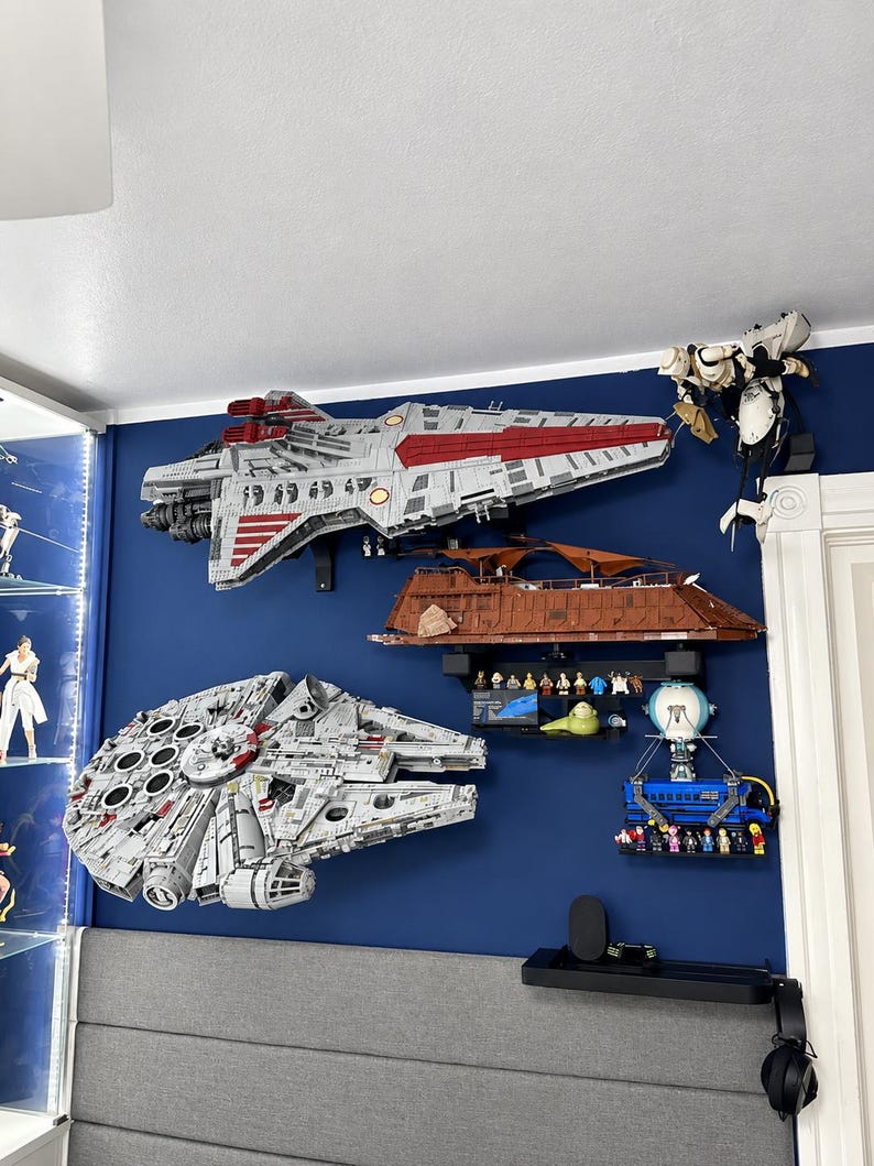Pu&ograve; includere: Una collezione di modelli costruibili a tema Star Wars montati su una parete blu scuro, tra cui una grande astronave grigia e rossa, un mercantile circolare grigio, un sandcrawler marrone, un veicolo blu e varie piccole figure.