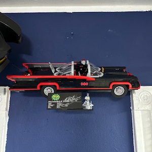 May include: A black Batmobile (1966) model with red trim and a clear windshield is mounted on a blue wall. The model has a sleek design with fins and is accompanied by a small sign and a Batman figure.