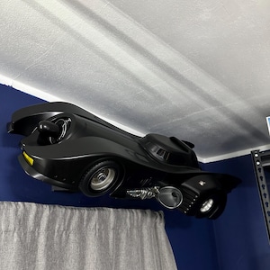 May include: A black Batmobile toy car with yellow accents, mounted on a wall. The toy car has detailed features, including wheels, exhaust pipes, and a cockpit. The car is positioned against a blue wall, with a gray curtain in the foreground.