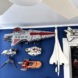 K&ouml;nnte beinhalten: Eine Sammlung von montierten Kunststoff-Bausteinmodellen, darunter ein gro&szlig;es graues und rotes Raumschiff, ein wei&szlig;er &Uuml;berschalljet, ein silberner Sportwagen, ein buntes Raumschiff, ein grauer J&auml;ger und ein orange-roter Gleiter an einer blauen Wand.