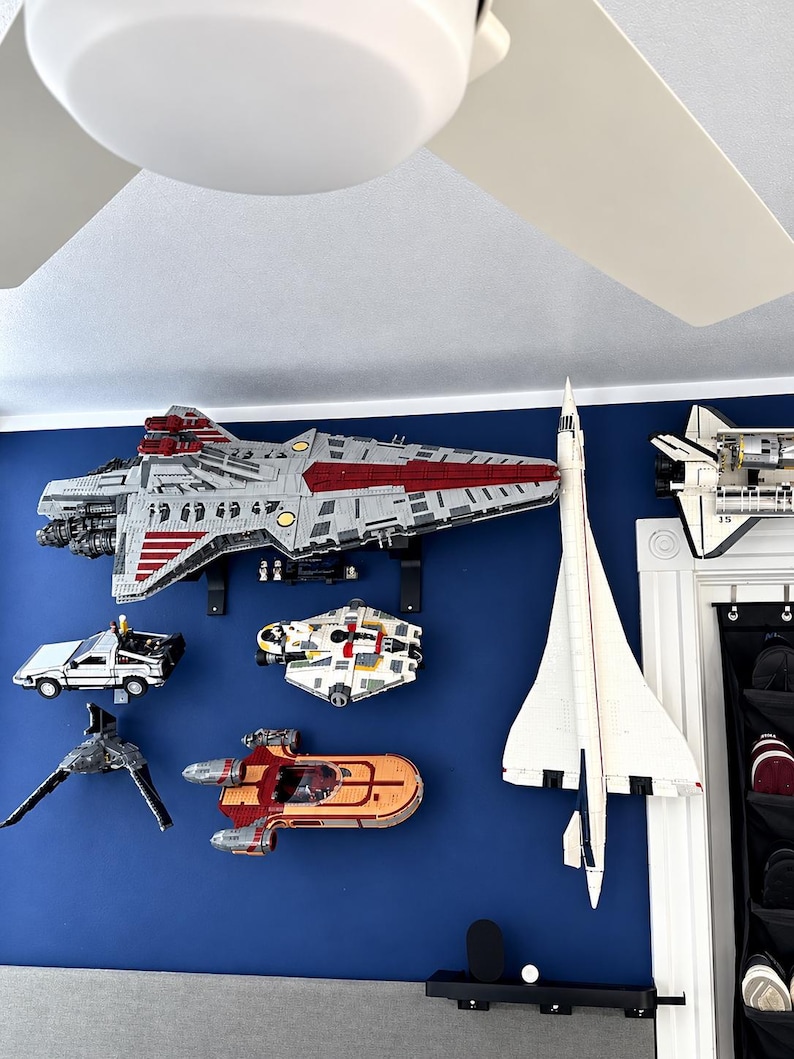 K&ouml;nnte beinhalten: Eine Sammlung von montierten Kunststoff-Bausteinmodellen, darunter ein gro&szlig;es graues und rotes Raumschiff, ein wei&szlig;er &Uuml;berschalljet, ein silberner Sportwagen, ein buntes Raumschiff, ein grauer J&auml;ger und ein orange-roter Gleiter an einer blauen Wand.
