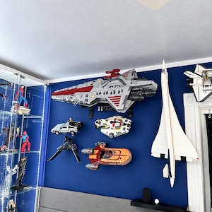 K&ouml;nnte beinhalten: Eine Sammlung von wandmontierten Modellraumschiffen und Flugzeugen, darunter ein gro&szlig;es graues und rotes Mutterschiff, eine wei&szlig;e Concorde, ein silberner Sportwagen, ein wei&szlig;-roter Sternenj&auml;ger und ein orangefarbener Landspeeder an einer blauen Wand.