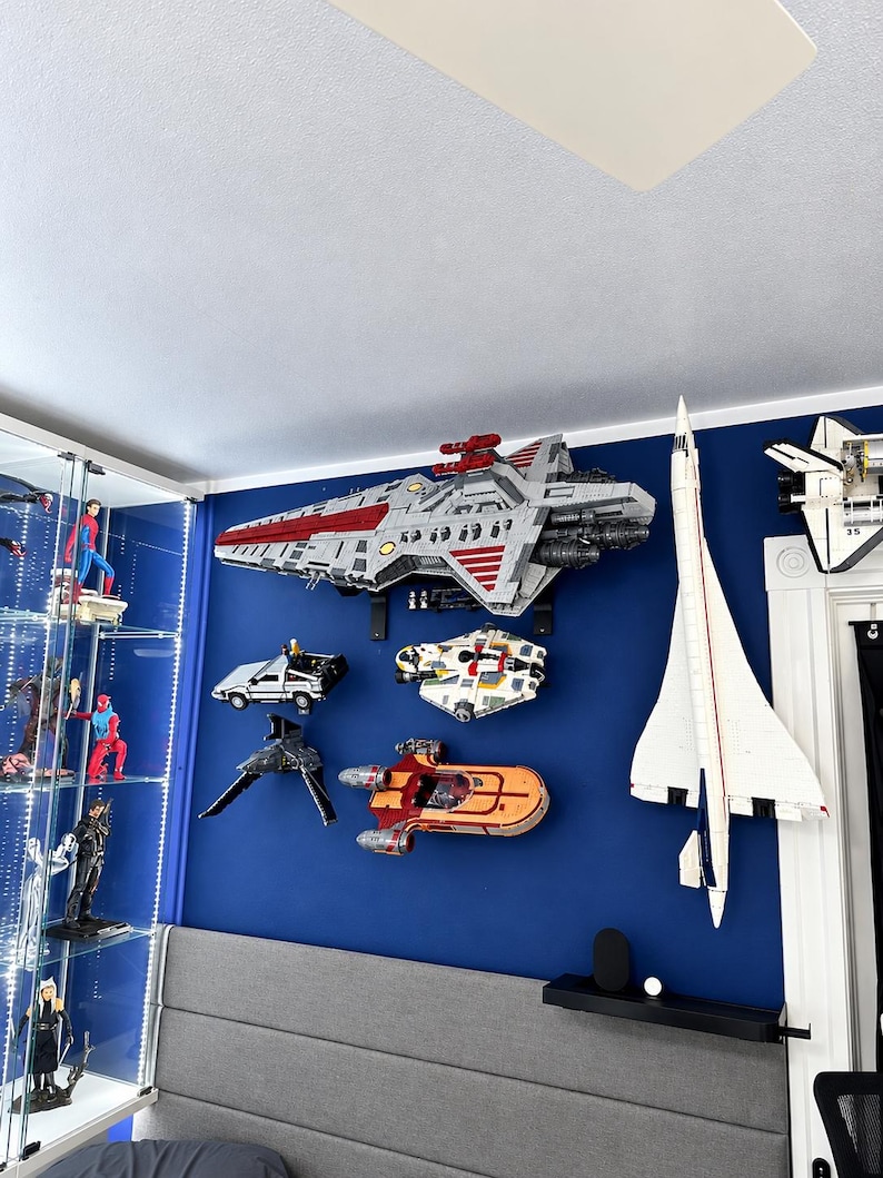 K&ouml;nnte beinhalten: Eine Sammlung von wandmontierten Modellraumschiffen und Flugzeugen, darunter ein gro&szlig;es graues und rotes Mutterschiff, eine wei&szlig;e Concorde, ein silberner Sportwagen, ein wei&szlig;-roter Sternenj&auml;ger und ein orangefarbener Landspeeder an einer blauen Wand.