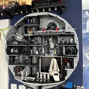 Op de afbeelding: Een gedetailleerd LEGO Death Star-model, met interieurniveaus met verschillende Star Wars-personages en -voertuigen. Het model is grijs met zwarte accenten, gemonteerd aan een muur. Bovenop staat een LEGO Hogwarts Express trein en station.