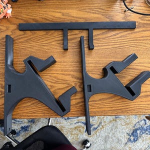 K&ouml;nnte beinhalten: Ein Set aus drei schwarzen 3D-gedruckten Komponenten, bestehend aus zwei identischen Wandhalterungen mit abgewinkelten St&uuml;tzarmen und einer horizontalen Querstange mit zwei nach unten gerichteten Haken, alle mit matter, texturierter Oberfl&auml;che.