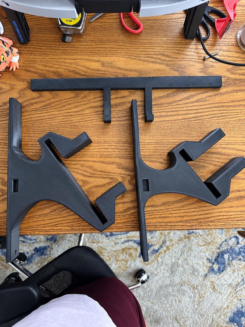 K&ouml;nnte beinhalten: Ein Set aus drei schwarzen 3D-gedruckten Komponenten, bestehend aus zwei identischen Wandhalterungen mit abgewinkelten St&uuml;tzarmen und einer horizontalen Querstange mit zwei nach unten gerichteten Haken, alle mit matter, texturierter Oberfl&auml;che.