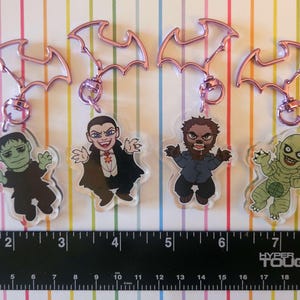 Puede incluir: Cuatro llaveros acrílicos con personajes de monstruos de dibujos animados: Frankenstein, Drácula, un hombre lobo y la Criatura de la Laguna Negra. Cada llavero tiene un cierre rosa en forma de murciélago y un anillo plateado.