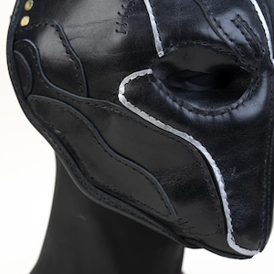 May include: A black leather mask with silver painted accents and decorative stitching. The mask has a contoured design with eye openings and is displayed on a black mannequin head. Two small gold rivets are visible.