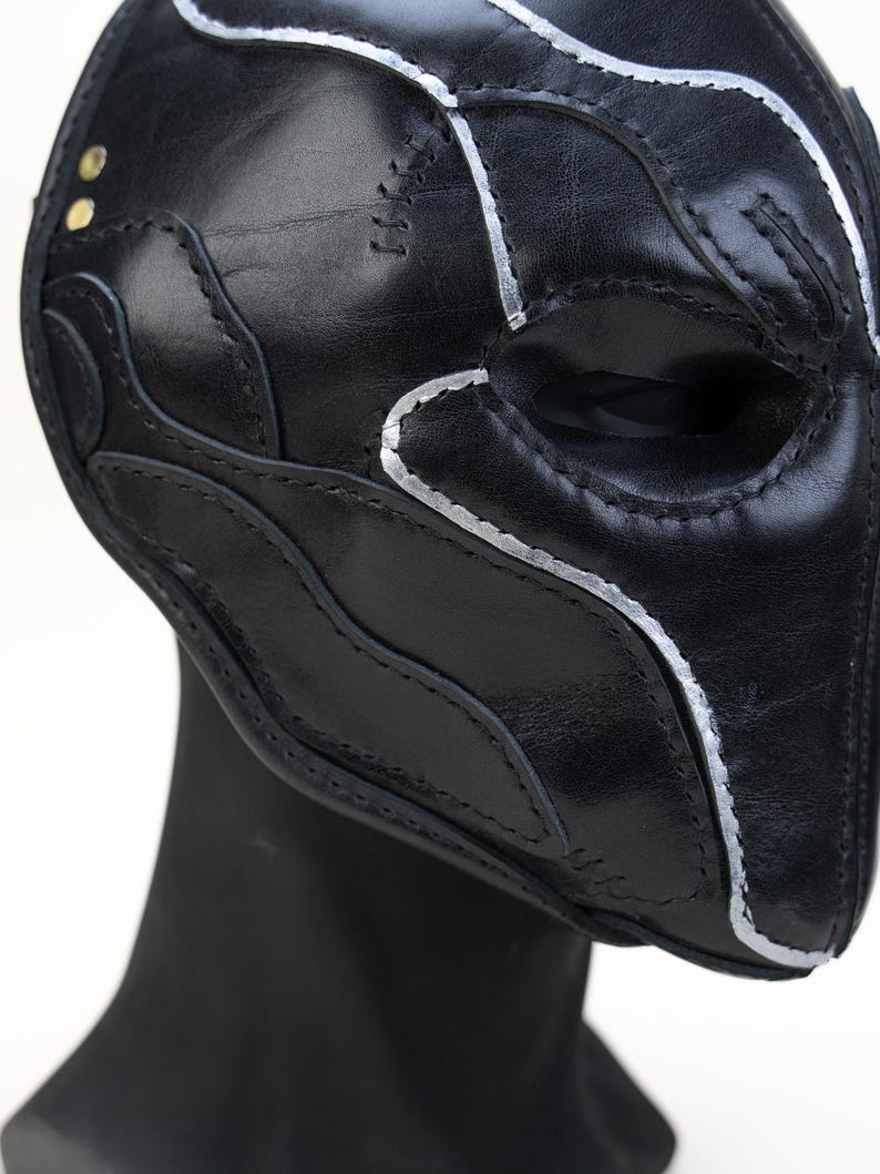 May include: A black leather mask with silver painted accents and decorative stitching. The mask has a contoured design with eye openings and is displayed on a black mannequin head. Two small gold rivets are visible.