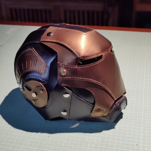 May include: A handcrafted leather helmet in shades of brown, black, and bronze. The helmet features riveted accents, a visor, and a detailed design, suggesting a futuristic or fantasy theme. The craftsmanship is evident in the stitching and panel construction.