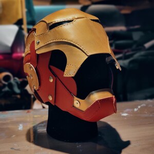 May include: A handcrafted, two-tone helmet in gold and red, likely for cosplay or display. The helmet features a detailed design with visible stitching and metallic accents. The color scheme and shape suggest a superhero or sci-fi theme.