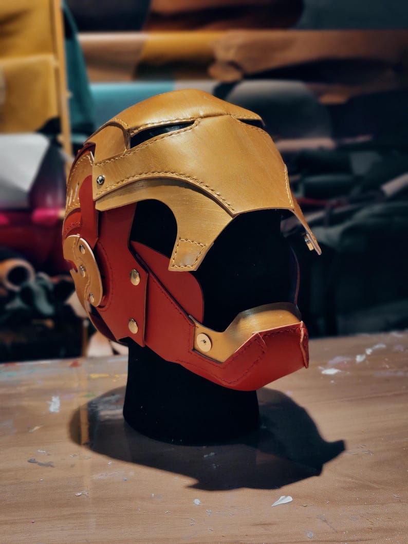 May include: A handcrafted, two-tone helmet in gold and red, likely for cosplay or display. The helmet features a detailed design with visible stitching and metallic accents. The color scheme and shape suggest a superhero or sci-fi theme.