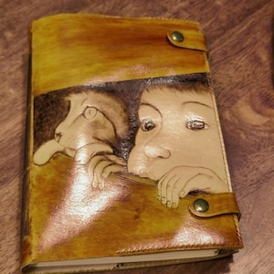 May include: Hand-tooled leather journal with a cat and child design. The cover is a warm, golden-brown colour with a darker brown illustration. The journal has a snap closure and appears to be new.