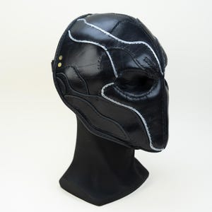 May include: A black leather mask with silver stitching and detailing. The mask is displayed on a black stand against a white background. The mask has a unique, sculpted design.
