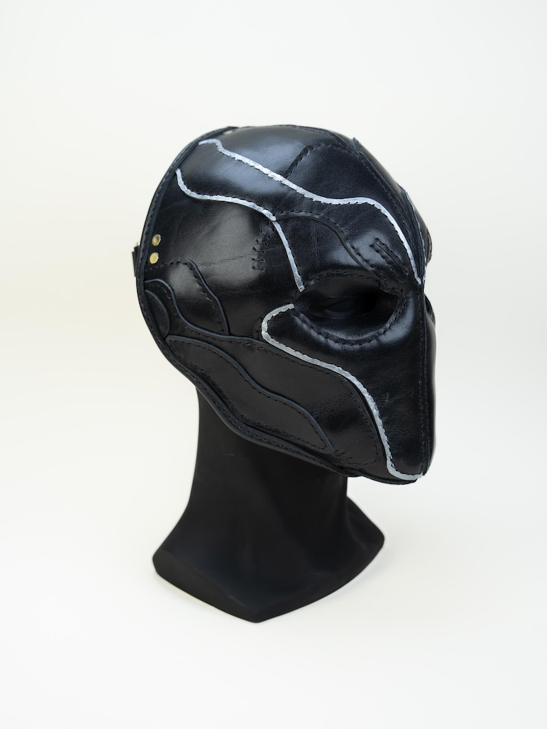 May include: A black leather mask with silver stitching and detailing. The mask is displayed on a black stand against a white background. The mask has a unique, sculpted design.