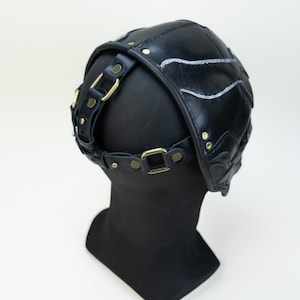 May include: Black leather head covering with decorative stitching and brass-colored hardware. The head covering is secured with a strap and buckle system. The design features a series of curved lines and is displayed on a black mannequin head.