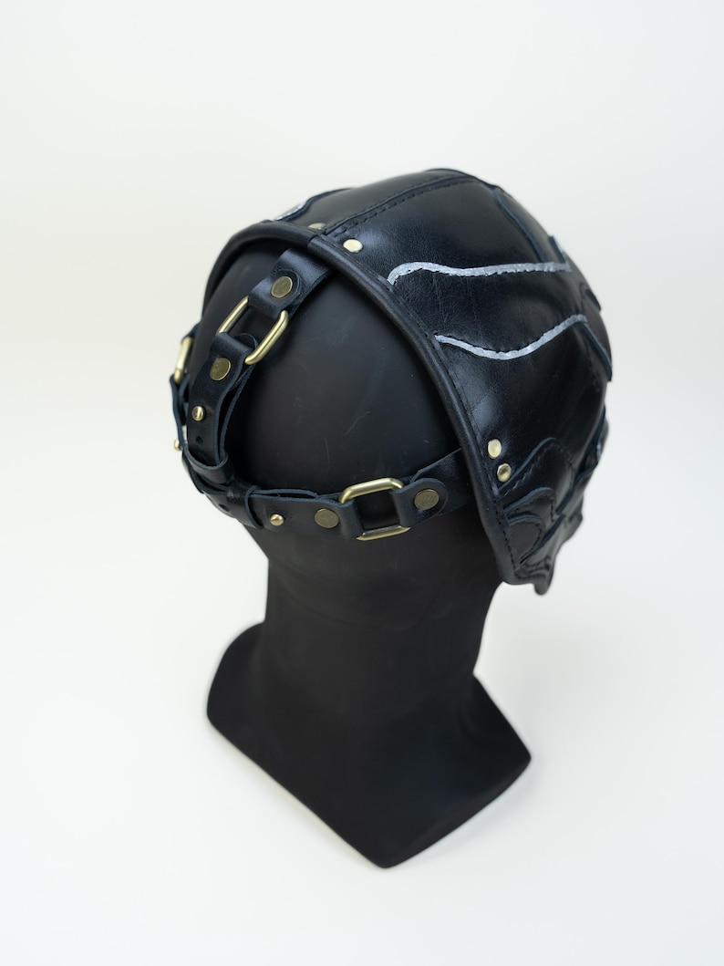 May include: Black leather head covering with decorative stitching and brass-colored hardware. The head covering is secured with a strap and buckle system. The design features a series of curved lines and is displayed on a black mannequin head.