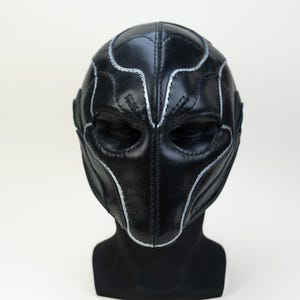 May include: A black leather mask with silver stitching. The mask has a symmetrical design with curved lines and shapes around the eyes and forehead. The mask is displayed on a black mannequin head against a white background.