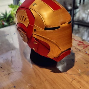 May include: A red and gold Iron Man helmet, featuring a detailed design with rivets and layered panels. The helmet is displayed on a black stand, showcasing the iconic superhero aesthetic. The color scheme includes a vibrant red and metallic gold.