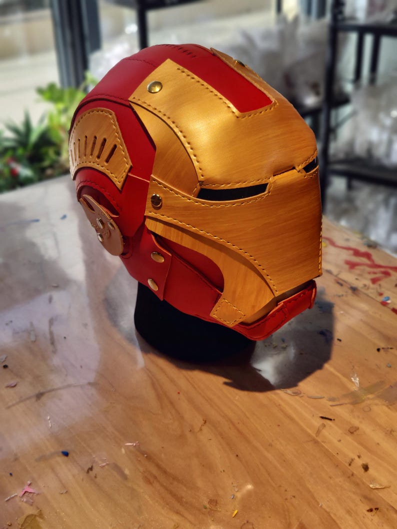 May include: A red and gold Iron Man helmet, featuring a detailed design with rivets and layered panels. The helmet is displayed on a black stand, showcasing the iconic superhero aesthetic. The color scheme includes a vibrant red and metallic gold.