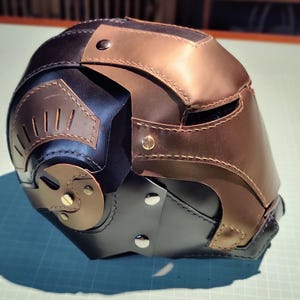 May include: A handcrafted leather helmet in black, bronze, and brown tones. The helmet features riveted accents, cut-out details, and a multi-panel design. The overall aesthetic suggests a futuristic or fantasy theme.