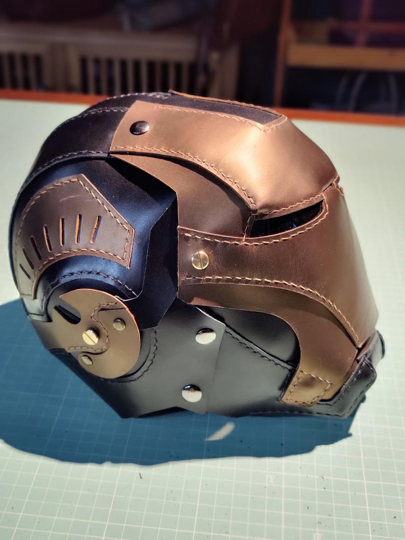 May include: A handcrafted leather helmet in black, bronze, and brown tones. The helmet features riveted accents, cut-out details, and a multi-panel design. The overall aesthetic suggests a futuristic or fantasy theme.