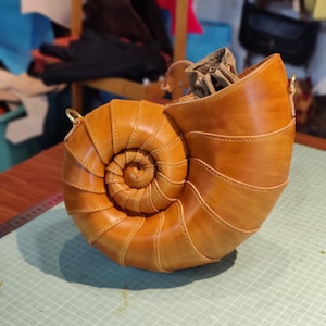 Könnte beinhalten: Handgefertigte, hellbraune Lederhandtasche in Form eines Nautilus-Gehäuses. Die Tasche hat ein Spiraldesign, sichtbare Nähte und einen Kordelzugverschluss. Sie hat einen Schulterriemen und goldfarbene Beschläge.