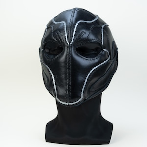 May include: A black leather mask with silver stitching and accents. The mask covers the eyes and nose, with openings for the eyes. The mask is displayed on a black mannequin head against a white background. The mask has a symmetrical design.