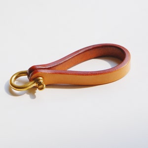 May include: A handcrafted leather keyring with a loop design. The keyring features a light brown leather strap with a darker brown edge and a brass-coloured metal clasp. The loop is approximately 10 cm long.