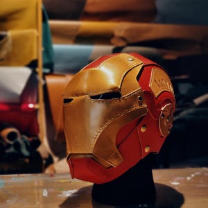 May include: A handcrafted red and gold leather helmet, reminiscent of a superhero's mask. The helmet features a gold faceplate, red accents, and decorative gold rivets. The design is detailed and appears to be a cosplay item.